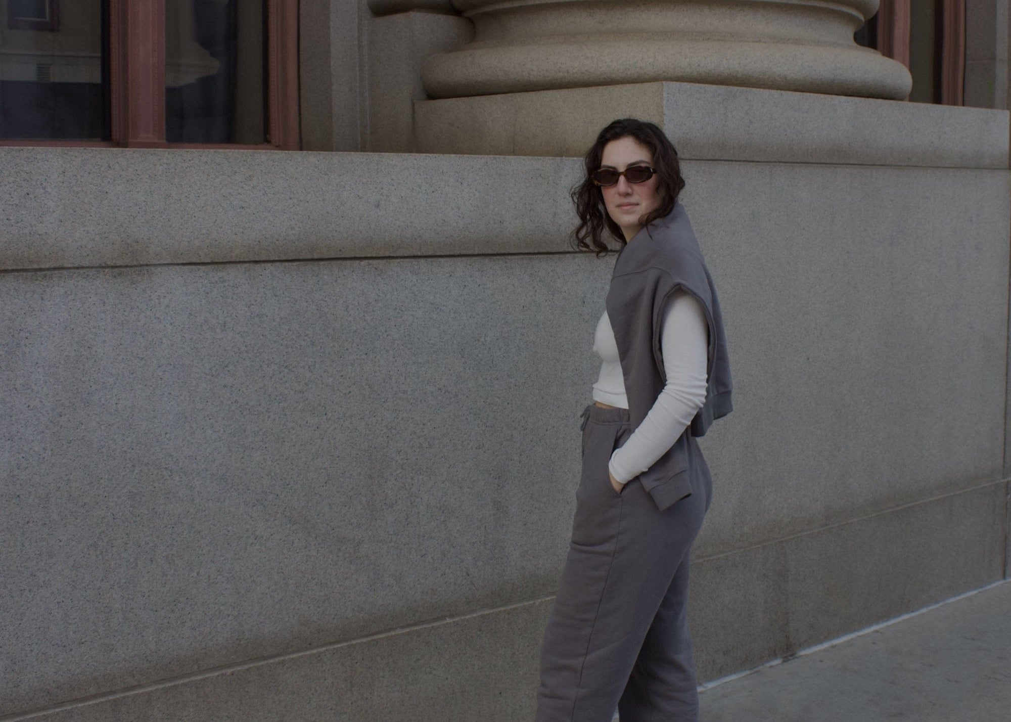Woman standing near a building with a sweatshirt draped over her shoulders and wearing grey sweatss